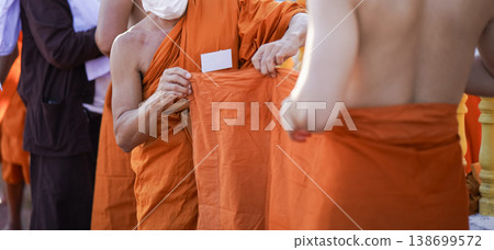 A senior monk adjusts a novice robe as hands tighten the woven belt around orange cloth in sunlight with focus on texture folds and precise fitting. A senior monk adjusts a novice robe as hands tighten the woven belt around orange cloth in sunlight with focus on texture folds and precise fitting. 138699572