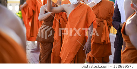 A senior monk adjusts a novice robe as hands tighten the woven belt around orange cloth in sunlight with focus on texture folds and precise fitting. 138699573