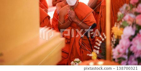 New monks sit in a large group inside the hall during ordination. Orange robes and warm temple light fill the frame. 138700321