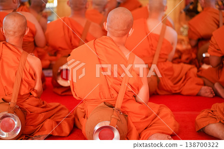 New monks sit in a large group inside the hall during ordination. Orange robes and warm temple light fill the frame. 138700322