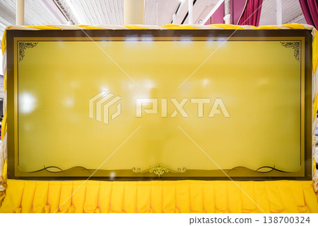A large blank yellow sign board stands indoors with a dark border. Decorative corners and draped fabric frame the panel. A large blank yellow sign board stands indoors with a dark border. Decorative corners and draped fabric frame the panel. 138700324
