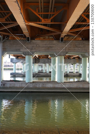 The orange-colored steel framework and concrete piers on the underside of the bridge are reflected on the water's surface. 138700600