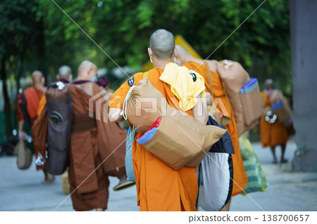 New monks walk from behind carrying supplies along a forest road. Orange robes and bags mark a quiet retreat journey. New monks walk from behind carrying supplies along a forest road. Orange robes and bags mark a quiet retreat journey. 138700657