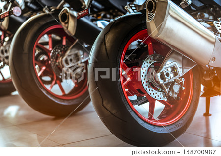 Closeup of new motorbike rear wheel. Big bike parked in showroom of dealership. Motorcycle exhaust pipes. Iconic motorcycle with sports design. Black tire with unique pattern and red spoked wheels. Closeup of new motorbike rear wheel. Big bike parked in showroom of dealership. Motorcycle exhaust pipes. Iconic motorcycle with sports design. Black tire with unique pattern and red spoked wheels. 138700987