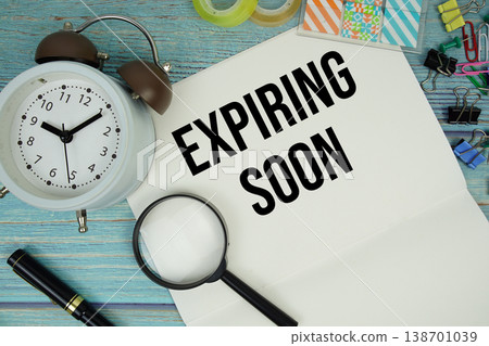 Expiring Soon text on a paper surrounded by office supplies, a magnifying glass, and a clock on a blue wooden surface 138701039