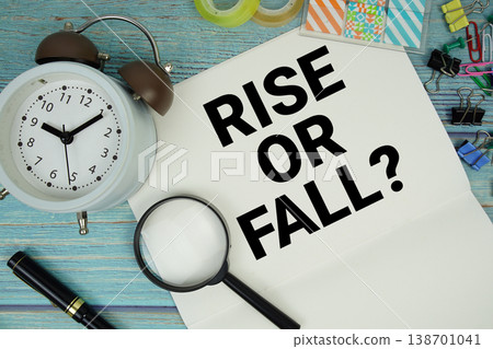 Rise Or Fall? text on a paper surrounded by office supplies, a magnifying glass, and a clock on a blue wooden surface 138701041