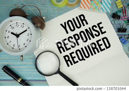 Your Response Required text on a paper surrounded by office supplies, a magnifying glass, and a clock on a blue wooden surface 138701044