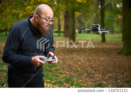 Man controlling flying drone. Bearded man operating quadcopter in park, focused on flight and reacting with excitement, concept of modern technology and hobby Man controlling flying drone. Bearded man operating quadcopter in park, focused on flight and reacting with excitement, concept of modern technology and hobby 138701131
