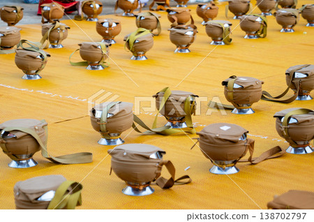 Dozens of covered alms bowls sit spaced across a yellow mat. Silver bases and fabric straps show an organized setup. 138702797