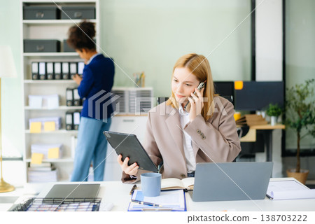 Young woman, smiling and working at her desk in an office environment. She is wearing a business suit Young woman, smiling and working at her desk in an office environment. She is wearing a business suit 138703222