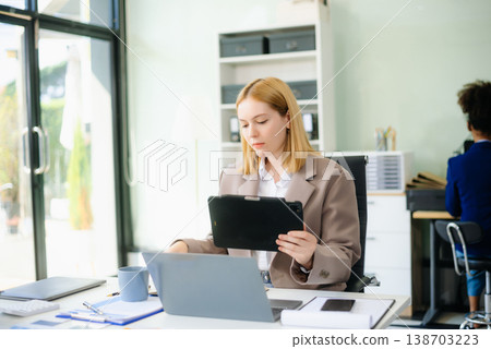 Young woman, smiling and working at her desk in an office environment. She is wearing a business suit Young woman, smiling and working at her desk in an office environment. She is wearing a business suit 138703223
