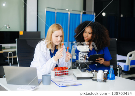 Focused Female doctor and African female nurse look at tablet and laptop screen discuss anamnesis together. Focused Female doctor and African female nurse look at tablet and laptop screen discuss anamnesis together. 138703454
