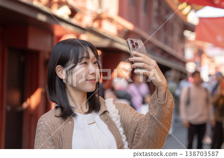 A smiling woman takes a selfie with her smartphone while walking through a downtown area. (Tourist spot, city walk, travel, lifestyle) A smiling woman takes a selfie with her smartphone while walking through a downtown area. (Tourist spot, city walk, travel, lifestyle) 138703587
