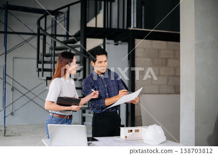 Asian architects collaborating with floor plans, tablet, and building models at a modern construction site. 138703812