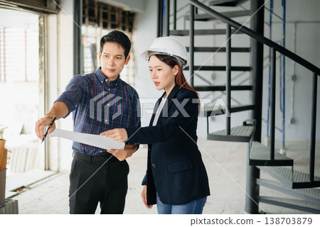 Asian engineers wearing hard hats discuss blueprints and floor plans at a construction site. Teamwork, planning, and innovation 138703879