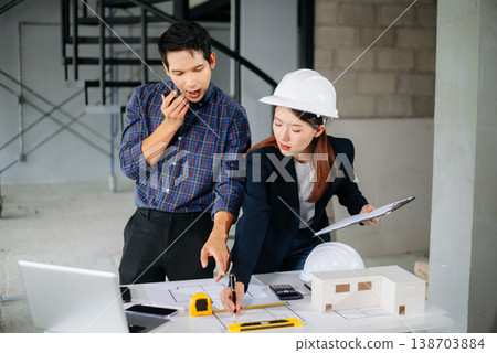 Asian engineers wearing hard hats discuss blueprints and floor plans at a construction site. Teamwork, planning, and innovation Asian engineers wearing hard hats discuss blueprints and floor plans at a construction site. Teamwork, planning, and innovation 138703884