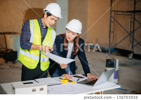 Asian architects at construction site using laptop and blueprints for project planning. Wearing hard hats and vests, they discuss design Asian architects at construction site using laptop and blueprints for project planning. Wearing hard hats and vests, they discuss design 138703938