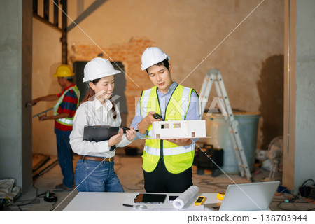 Asian engineers at building site reviewing architecture model. Wearing hard hats and safety vests, they discuss layout, planning, teamwork Asian engineers at building site reviewing architecture model. Wearing hard hats and safety vests, they discuss layout, planning, teamwork 138703944