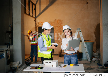 Asian engineers discuss floor plan using laptop and building model at active construction site. Collaboration, safety Asian engineers discuss floor plan using laptop and building model at active construction site. Collaboration, safety 138703952