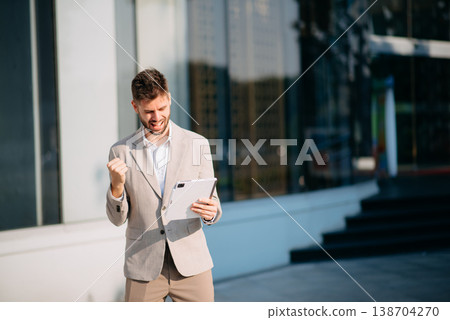 businessman works outdoors near the office holds a tablet, smiles reads good news businessman works outdoors near the office holds a tablet, smiles reads good news 138704270