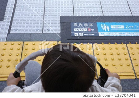 The image shows the back of a child in a stroller waiting for a train on a station platform, along with priority seating signs and tactile paving. The image shows the back of a child in a stroller waiting for a train on a station platform, along with priority seating signs and tactile paving. 138704341