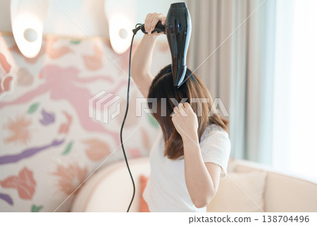 Young woman drying long hair with a hair dryer after a shower in home or hotel room during vacation. Hairstyles and daily morning routine Young woman drying long hair with a hair dryer after a shower in home or hotel room during vacation. Hairstyles and daily morning routine 138704496