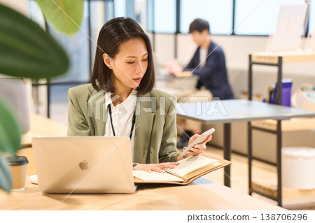 一位女系統工程師在外資IT公司的辦公室一邊看著智慧型手機一邊做筆記。 一位女系統工程師在外資IT公司的辦公室一邊看著智慧型手機一邊做筆記。 138706296