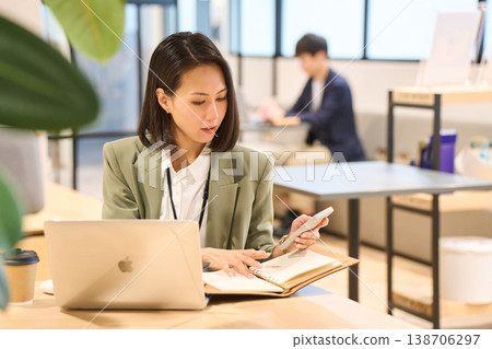 A female systems engineer taking notes while looking at her smartphone in the office of a foreign-owned IT company. A female systems engineer taking notes while looking at her smartphone in the office of a foreign-owned IT company. 138706297
