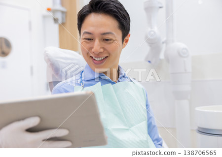 一位男患者在牙醫診所一邊聽著醫生給他看平板電腦,一邊聽著他的檢查結果。 一位男患者在牙醫診所一邊聽著醫生給他看平板電腦,一邊聽著他的檢查結果。 138706565