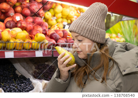 Woman smelling ripe pear in supermarket produce section. Shopper examining fresh fruit quality with grapes and apples Woman smelling ripe pear in supermarket produce section. Shopper examining fresh fruit quality with grapes and apples 138708540