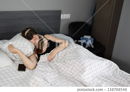 Woman lying relaxed in bed with black sleep mask over eyes and smartphone in hand. Tissues on nightstand in cozy bedroom setting. Restful sleep routine and self-care concept for wellness and Woman lying relaxed in bed with black sleep mask over eyes and smartphone in hand. Tissues on nightstand in cozy bedroom setting. Restful sleep routine and self-care concept for wellness and 138708548