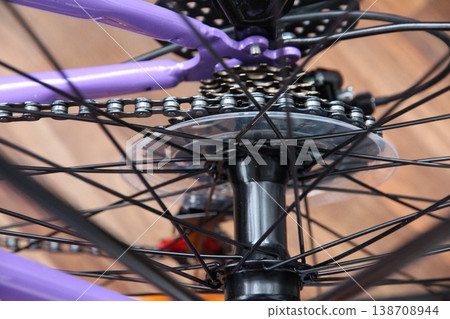 Close-up of a bicycle rear section with chain, spokes, and hub on a purple metal frame, indoors on a wooden floor, highlighting mechanical details, motion, and cycling lifestyle 138708944