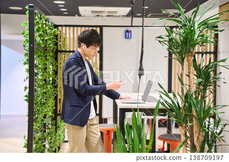 一位來自外國公司的年輕男性系統工程師在綠樹成蔭的辦公室裡,使用站立式辦公桌進行網路會議。 一位來自外國公司的年輕男性系統工程師在綠樹成蔭的辦公室裡,使用站立式辦公桌進行網路會議。 138709197