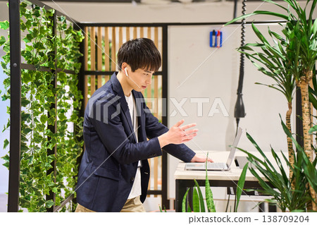 一位來自外國公司的年輕男性系統工程師在綠樹成蔭的辦公室裡,使用站立式辦公桌進行網路會議。 一位來自外國公司的年輕男性系統工程師在綠樹成蔭的辦公室裡,使用站立式辦公桌進行網路會議。 138709204