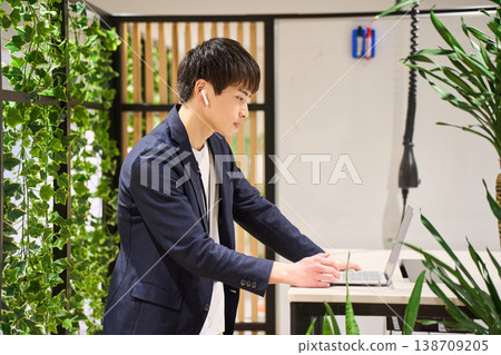 一位來自外國公司的年輕男性系統工程師在綠樹成蔭的辦公室裡,使用站立式辦公桌進行網路會議。 一位來自外國公司的年輕男性系統工程師在綠樹成蔭的辦公室裡,使用站立式辦公桌進行網路會議。 138709205
