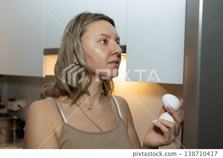 Woman holding two fresh eggs in modern kitchen near refrigerator. Morning breakfast preparation and healthy eating routine concept for daily nutrition and family meals. 138710417