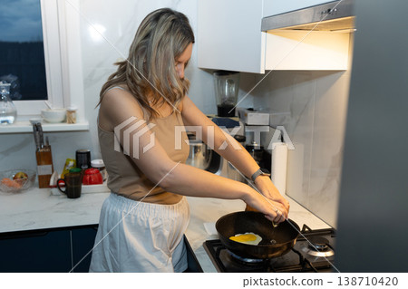 Woman frying egg on stove in modern kitchen. Morning breakfast preparation and daily routine. Fresh home cooking concept for healthy start to day. Woman frying egg on stove in modern kitchen. Morning breakfast preparation and daily routine. Fresh home cooking concept for healthy start to day. 138710420
