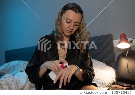 Thoughtful blonde woman in silk robe applies lotion to hand on bed photography. Thoughtful blonde woman in silk robe applies lotion to hand on bed photography. 138710435