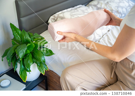 Woman holding ergonomic orthopedic pillow near bed preparing for sleep. Cervical spine support for proper neck alignment, posture correction and relief from tension, discomfort and muscle stiffness. Woman holding ergonomic orthopedic pillow near bed preparing for sleep. Cervical spine support for proper neck alignment, posture correction and relief from tension, discomfort and muscle stiffness. 138710846