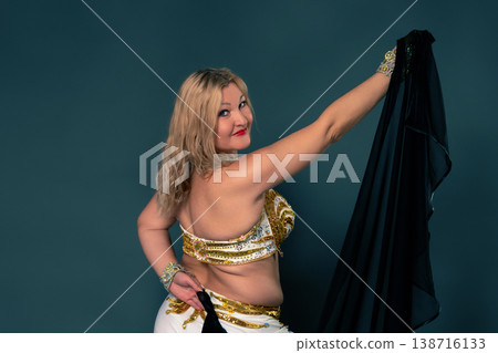 Woman dancer in white and golden belly dancing costume, looking over her shoulder, posing gracefully. She holding black veil against teal background. Studio shot with expressing culture and art 138716133
