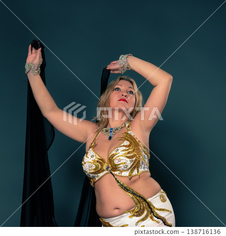 Graceful middle aged woman performs belly dance in an ornate white and golden costume with jewels. Dancer holds black veil against teal studio background. Evokes culture, art and performance Graceful middle aged woman performs belly dance in an ornate white and golden costume with jewels. Dancer holds black veil against teal studio background. Evokes culture, art and performance 138716136