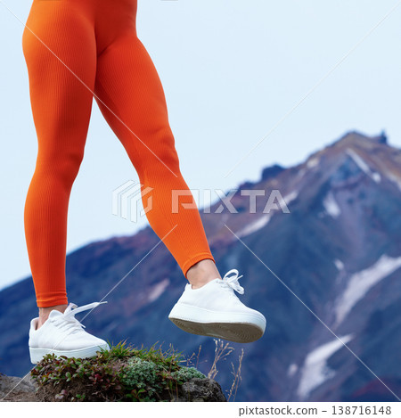 Close up of an athletic woman's legs in bright orange leggings and white sneakers. Female is hiking on a rocky peak with a scenic mountain view. Active, adventurous lifestyle concept 138716148