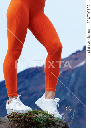 Low angle closeup of a fit woman's legs in bright orange ribbed leggings white sneakers. She stands on a rocky surface with mountains in the background. Concept of fitness, hiking, adventure Low angle closeup of a fit woman's legs in bright orange ribbed leggings white sneakers. She stands on a rocky surface with mountains in the background. Concept of fitness, hiking, adventure 138716152