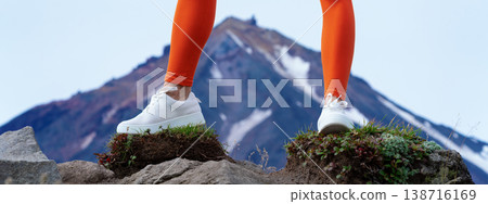 Low angle view of a fit woman's legs in bright orange leggings and white sneakers. She stands on a rocky summit with a snowy mountain in the background. Active lifestyle and travel concept Low angle view of a fit woman's legs in bright orange leggings and white sneakers. She stands on a rocky summit with a snowy mountain in the background. Active lifestyle and travel concept 138716169