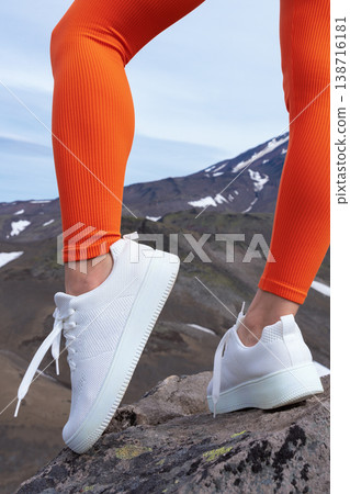 Closeup of a woman's legs in vibrant orange ribbed leggings and white sneakers. Female person stands on a rock, hiking in a mountain range. Active lifestyle concept with bold colors Closeup of a woman's legs in vibrant orange ribbed leggings and white sneakers. Female person stands on a rock, hiking in a mountain range. Active lifestyle concept with bold colors 138716181
