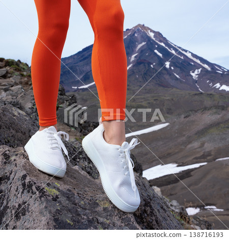 Low angle view of female legs in vibrant orange leggings and clean white sneakers. Standing on rocks in a rugged mountain landscape. Energetic and adventurous mood for fitness and travel 138716193