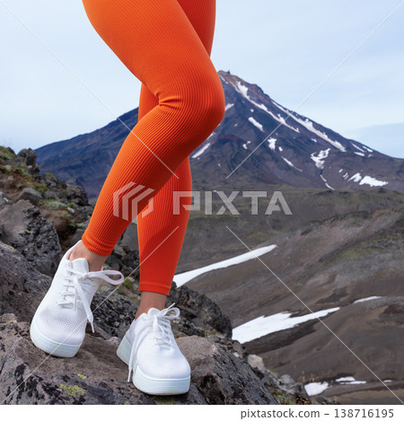 Close up of a fit woman's legs in bright orange ribbed leggings and white sneakers. She stands on a rocky mountain trail, with volcanic peak in the background. Active lifestyle concept Close up of a fit woman's legs in bright orange ribbed leggings and white sneakers. She stands on a rocky mountain trail, with volcanic peak in the background. Active lifestyle concept 138716195