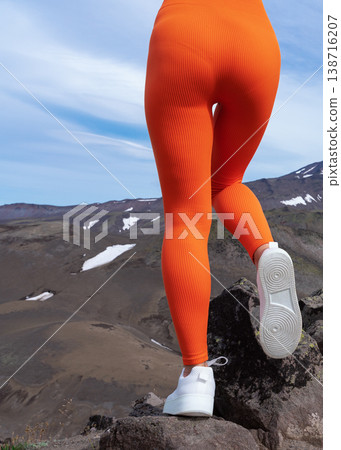 Low angle view of a fit woman's legs in vibrant orange ribbed leggings and white sneakers. Woman stands on a rocky summit, exploring a rugged mountain valley. Active lifestyle and travel concept Low angle view of a fit woman's legs in vibrant orange ribbed leggings and white sneakers. Woman stands on a rocky summit, exploring a rugged mountain valley. Active lifestyle and travel concept 138716207