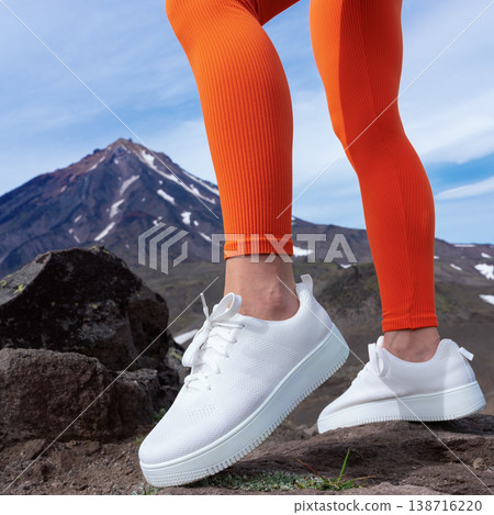 Close-up of a woman's legs in bright orange leggings and white sneakers on a rocky trail. A snow-capped mountain is in the background under a blue sky. Energetic, active lifestyle concept 138716220