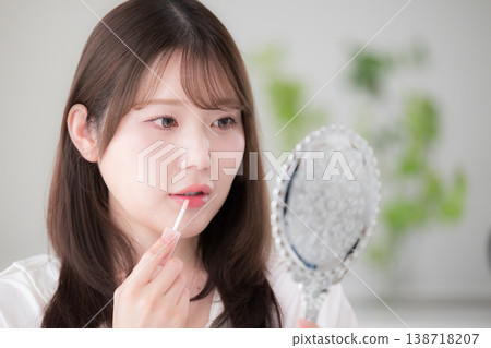 A woman applies lipstick while looking in a hand mirror, showcasing her natural makeup and beauty care routine, including lip gloss and lip care. 138718207